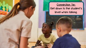 This photo shows a teacher sitting on the ground with 2 students talking with them.